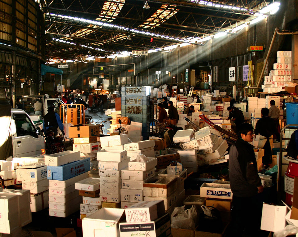 A large amount over boxes sit on top of each other in a chaotic manner in a Japanese warehouse.  The warehouse has sunlight shining through openings near the ceiling.
